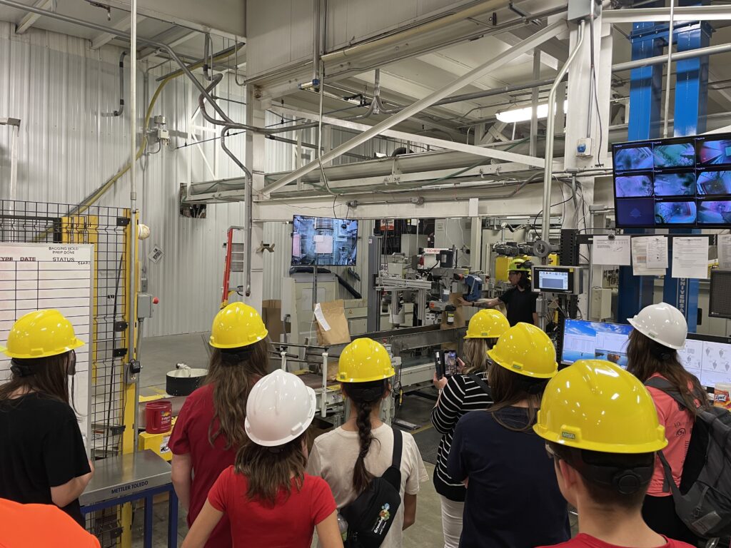 Students in hard hats watch a tour guide explaining equipment in an industry lab.